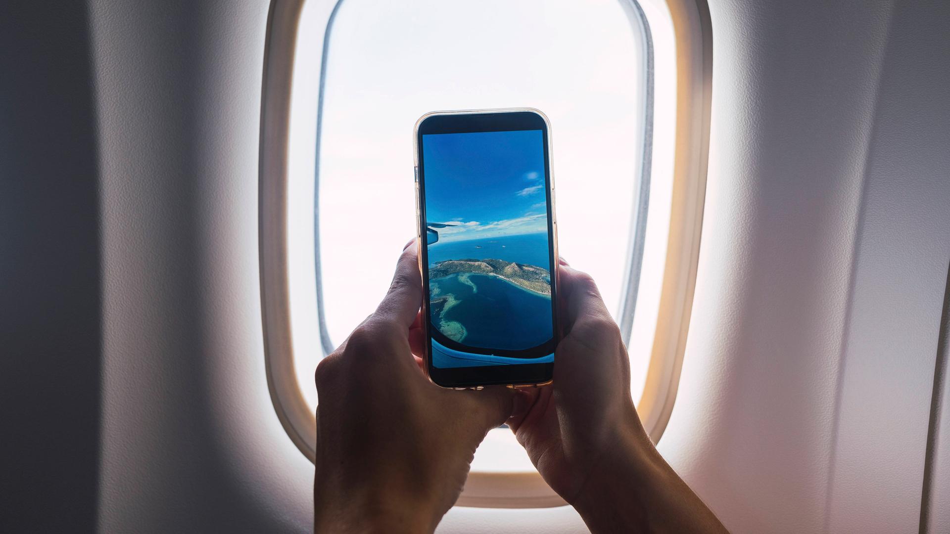 Ein Mann, von dem man nur die Hände sieht, macht mit dem Smartphone ein Foto durch das Fenster eines Flugzeugs.