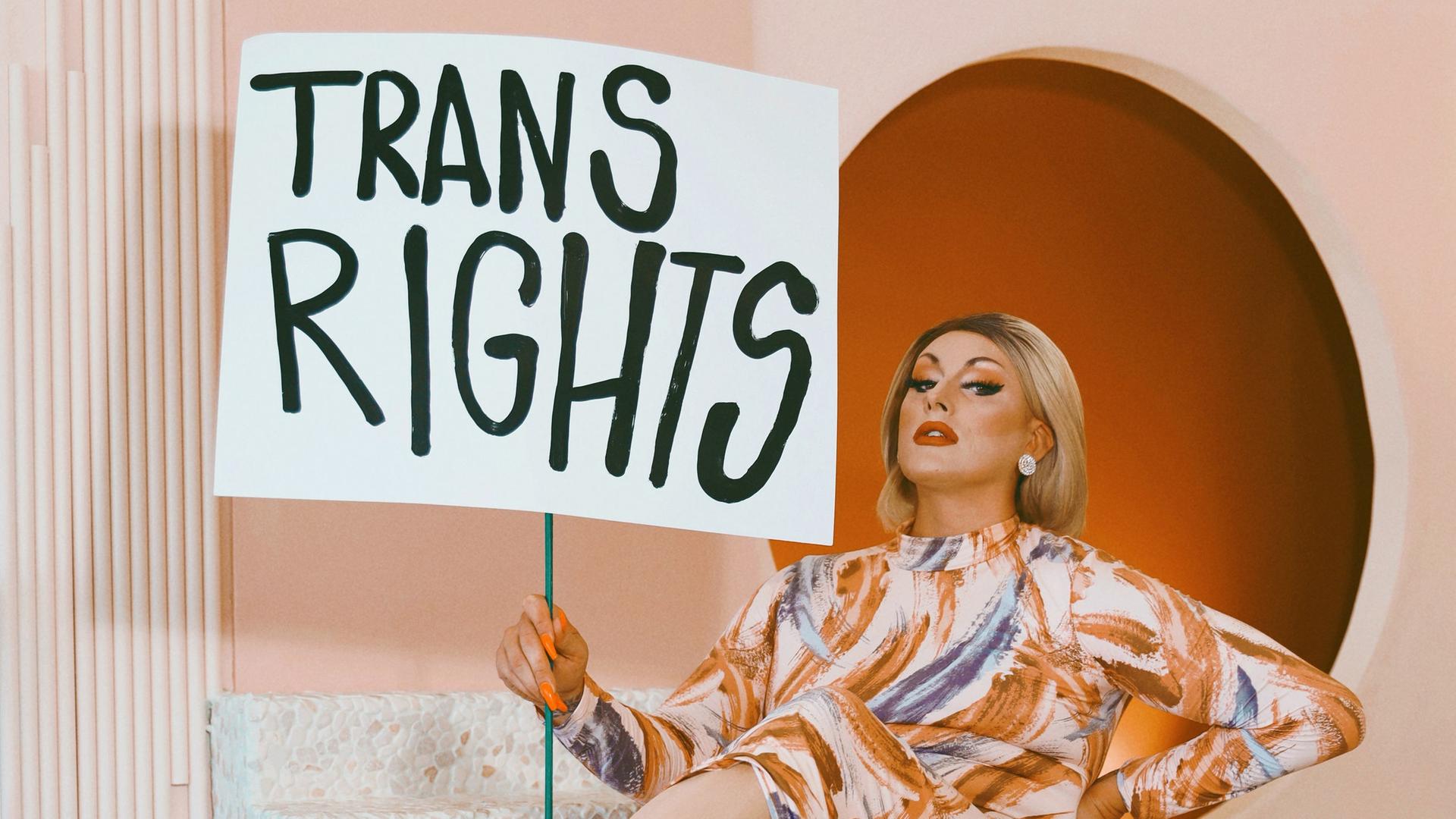 Die Dragqueen und Politikerin Maebe A. Girl mit einem Schild auf dem "Trans Rights" steht vor einer orangefarbenen Kulisse.