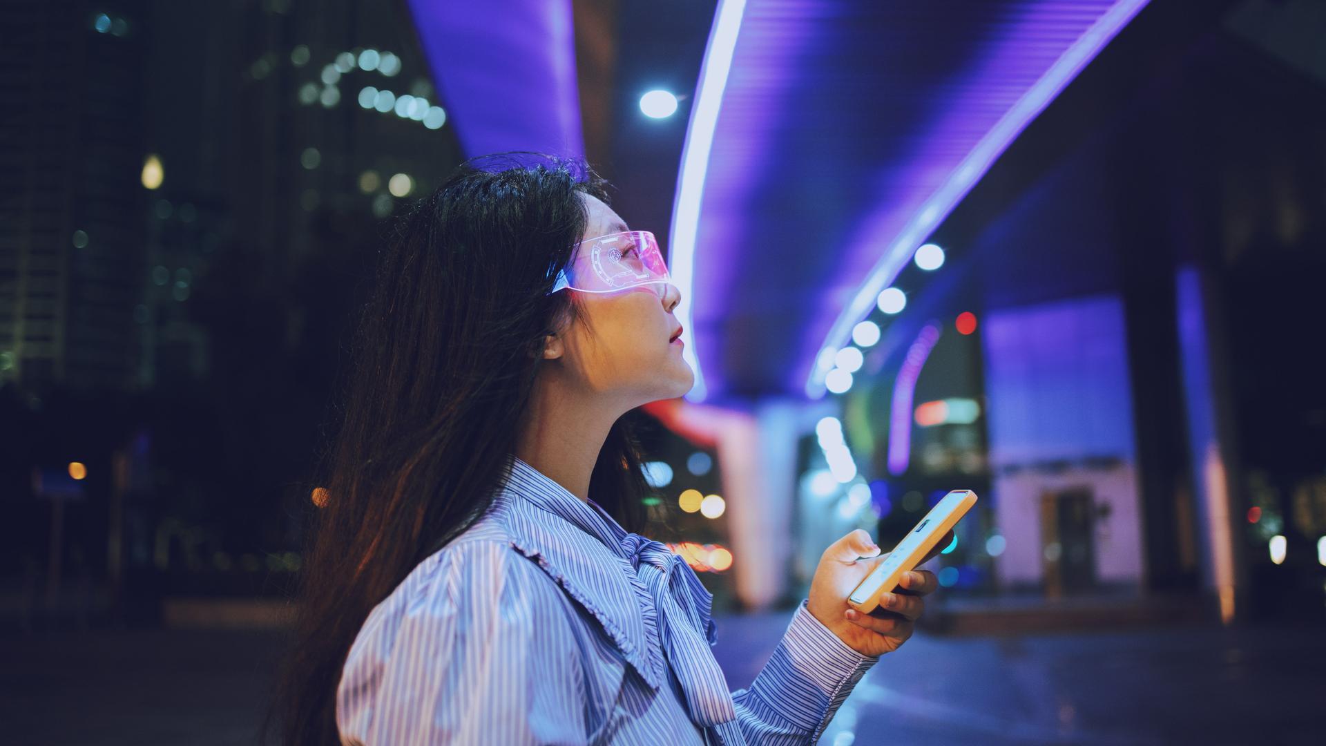 Eine junge Frau steht unter einer Brücke und nutz allemöglichen technischen Geräte. Eine Datenbrille und eine Art Smartphone. Im Hintergrund sind die Lichter der Stadt bei Nacht zu erkennen. Die Brücke unter der sie steht ist blau erleuchtet.