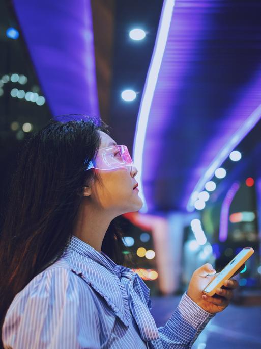 Eine junge Frau steht unter einer Brücke und nutz allemöglichen technischen Geräte. Eine Datenbrille und eine Art Smartphone. Im Hintergrund sind die Lichter der Stadt bei Nacht zu erkennen. Die Brücke unter der sie steht ist blau erleuchtet.