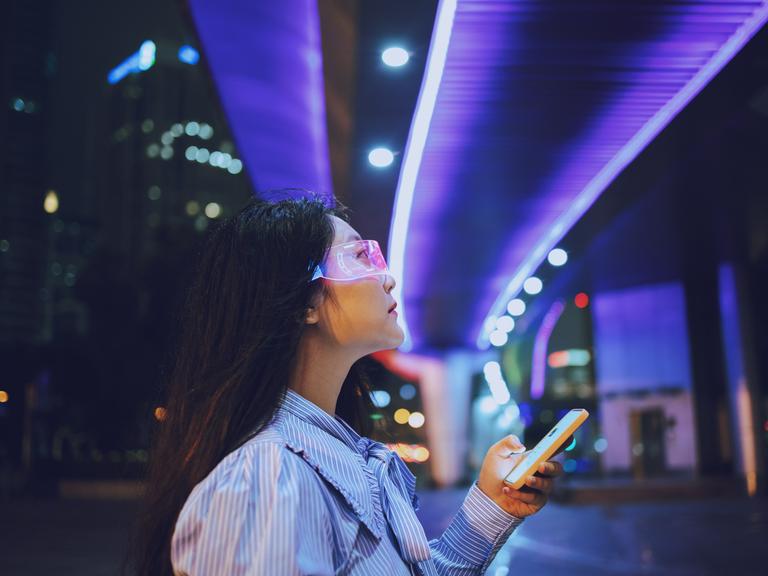 Eine junge Frau steht unter einer Brücke und nutz allemöglichen technischen Geräte. Eine Datenbrille und eine Art Smartphone. Im Hintergrund sind die Lichter der Stadt bei Nacht zu erkennen. Die Brücke unter der sie steht ist blau erleuchtet.