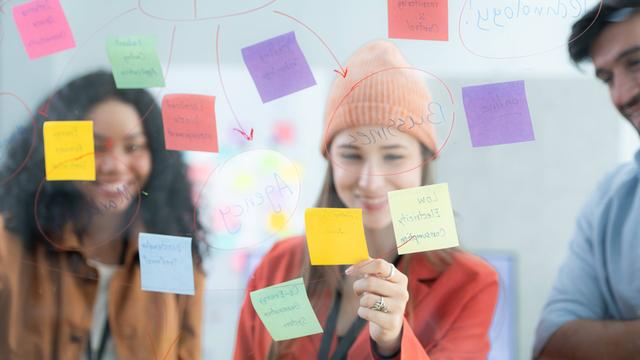 Ein Team von jungen Menschen, die Post-its auf einem transparenten Board anschauen. 