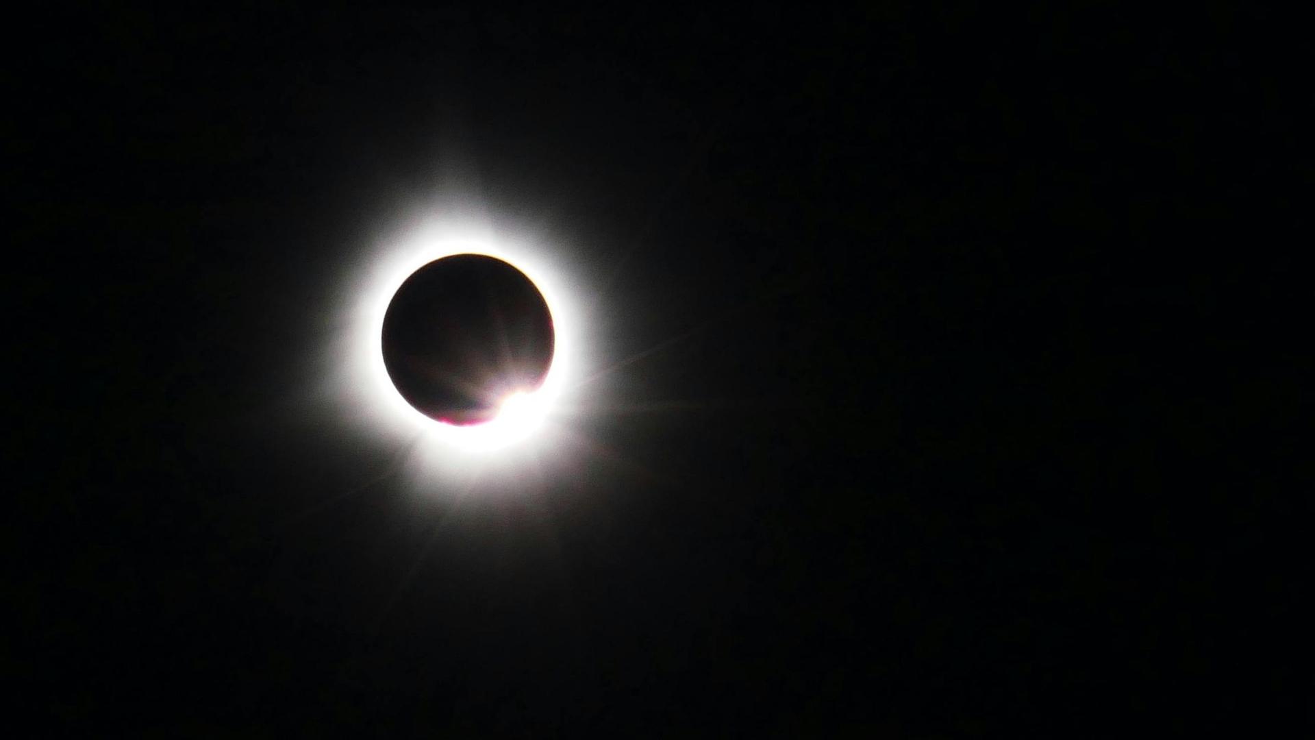 Blick auf eine Sonnenfinsternis, bei der der schwarze Mond die Sonne komplett bedeckt und nur ihre Strahlen um den schwarzen Punkt erscheinen.