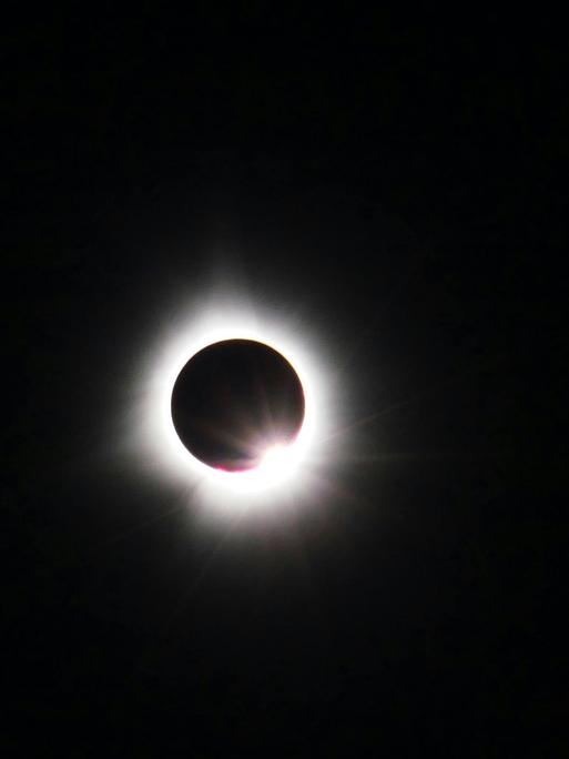 Blick auf eine Sonnenfinsternis, bei der der schwarze Mond die Sonne komplett bedeckt und nur ihre Strahlen um den schwarzen Punkt erscheinen.