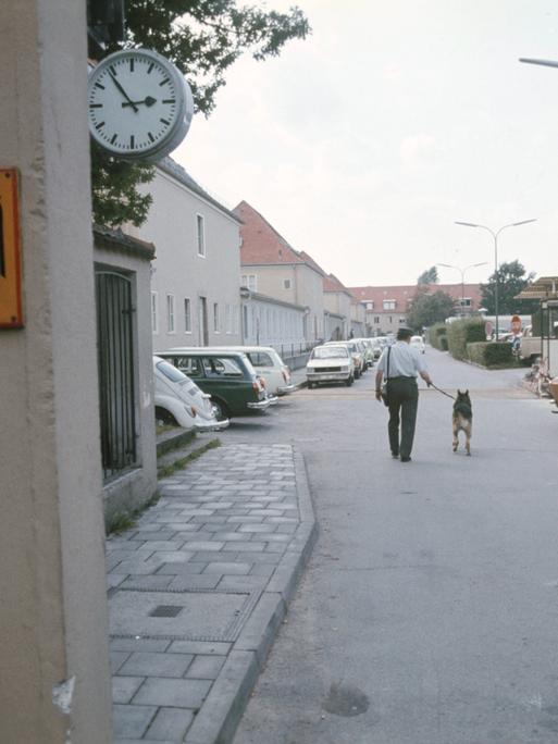 Durch ein Eingangstor sieht man einen Polizeibeamten mit Hund von hinten auf einer Aufnahme des Gelände des Bundesnachrichtendienstes in Pullach um 1960/1970.