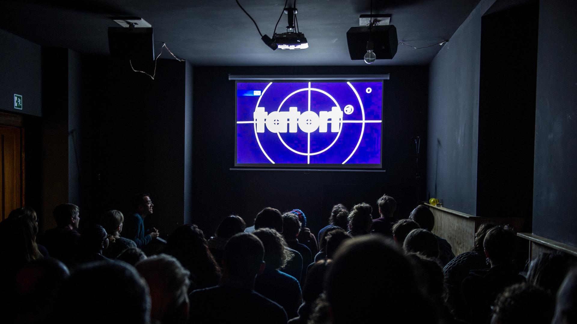 „Tatort“ ist für viele Menschen ein sonntägliches Ritual. Wie hier, wird auch oft zusammen in der Kneipe geguckt. Menschen sitzen in Stuhlreihen vor einer Leinwand auf der der „Tatort“ übertragen wird. „Tatort“ ist für viele Menschen ein sonntägliches Ritual. Wie hier, wird auch oft zusammen in der Kneipe geguckt. Menschen sitzen in Stuhlreihen vor einer Leinwand auf der der „Tatort“ übertragen wird.