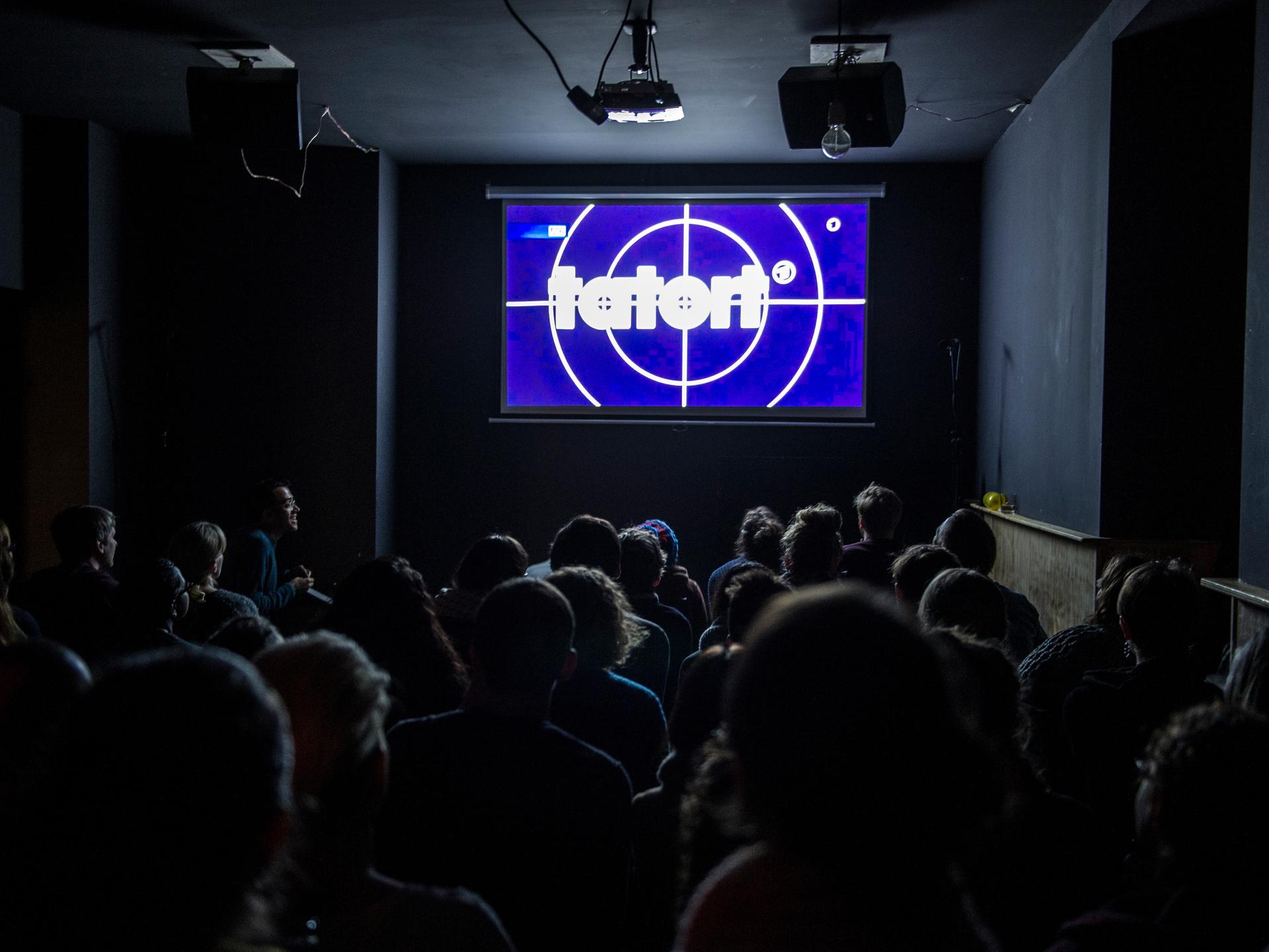 „Tatort“ ist für viele Menschen ein sonntägliches Ritual. Wie hier, wird auch oft zusammen in der Kneipe geguckt. Menschen sitzen in Stuhlreihen vor einer Leinwand auf der der „Tatort“ übertragen wird.
