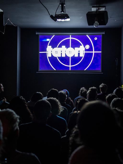 „Tatort“ ist für viele Menschen ein sonntägliches Ritual. Wie hier, wird auch oft zusammen in der Kneipe geguckt. Menschen sitzen in Stuhlreihen vor einer Leinwand auf der der „Tatort“ übertragen wird.