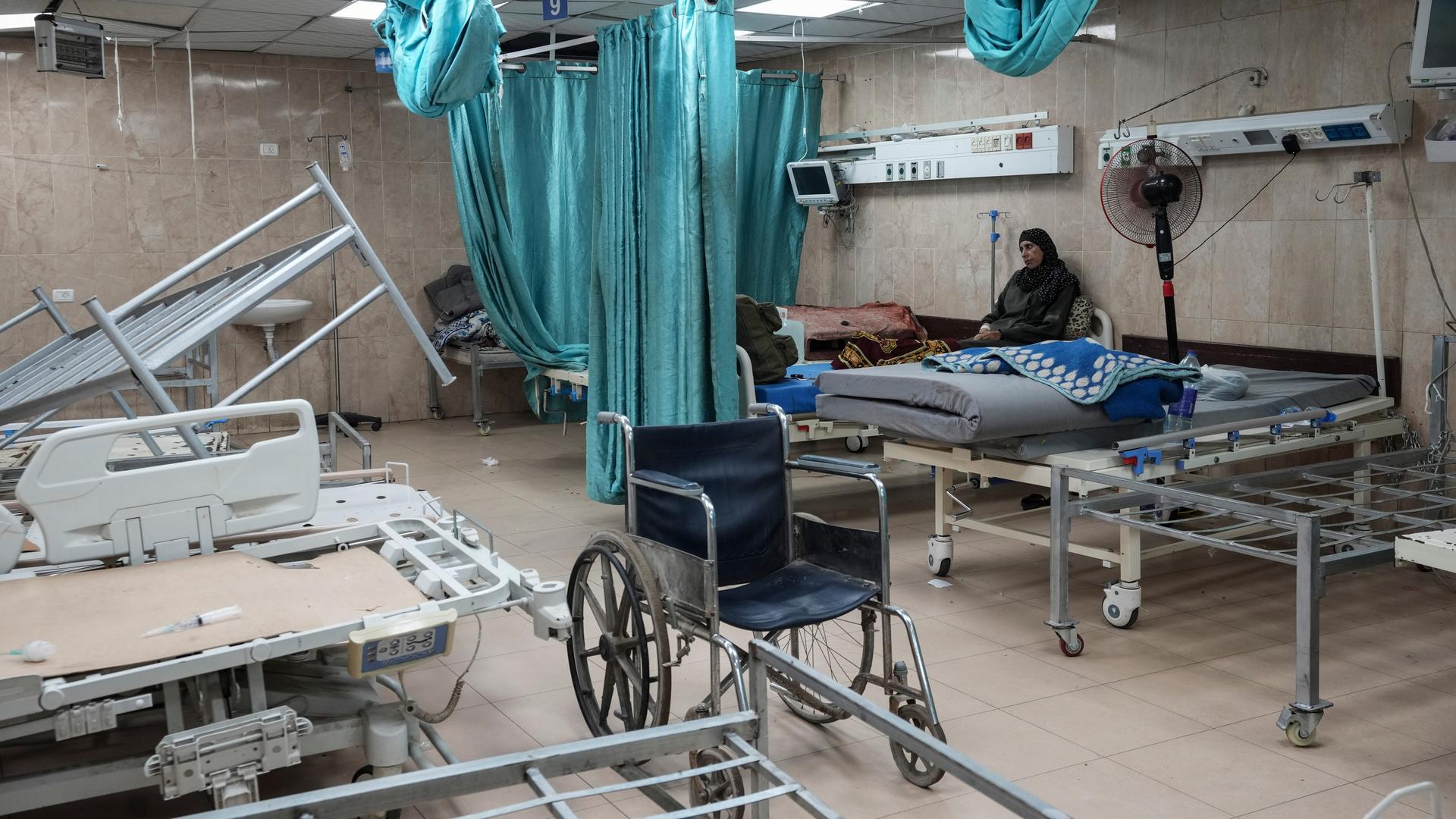 Eine arabisch aussehende Frau sitzt allein auf einem Bett in einem Krankenhauszimmer. Das Zimmer sieht verwüstet aus. Auf den meisten Betten sind keine Matratzen, ein Gestell liegt quer im Raum. Eine arabisch aussehende Frau sitzt allein auf einem Bett in einem Krankenhauszimmer. Das Zimmer sieht verwüstet aus. Auf den meisten Betten sind keine Matratzen, ein Gestell liegt quer im Raum.