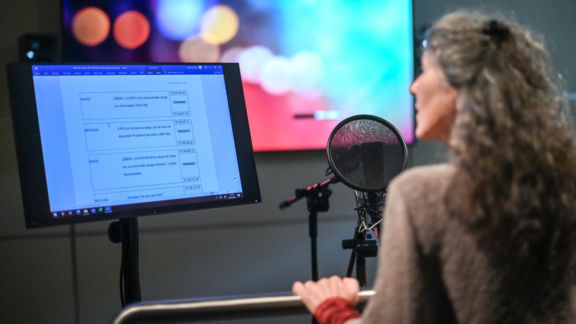 Eine Frau steht in einem Synchronstudio hinter einem Mikrofon und einem Bildschirm, der Dialoge anzeigt. Eine Frau steht in einem Synchronstudio hinter einem Mikrofon und einem Bildschirm, der Dialoge anzeigt.