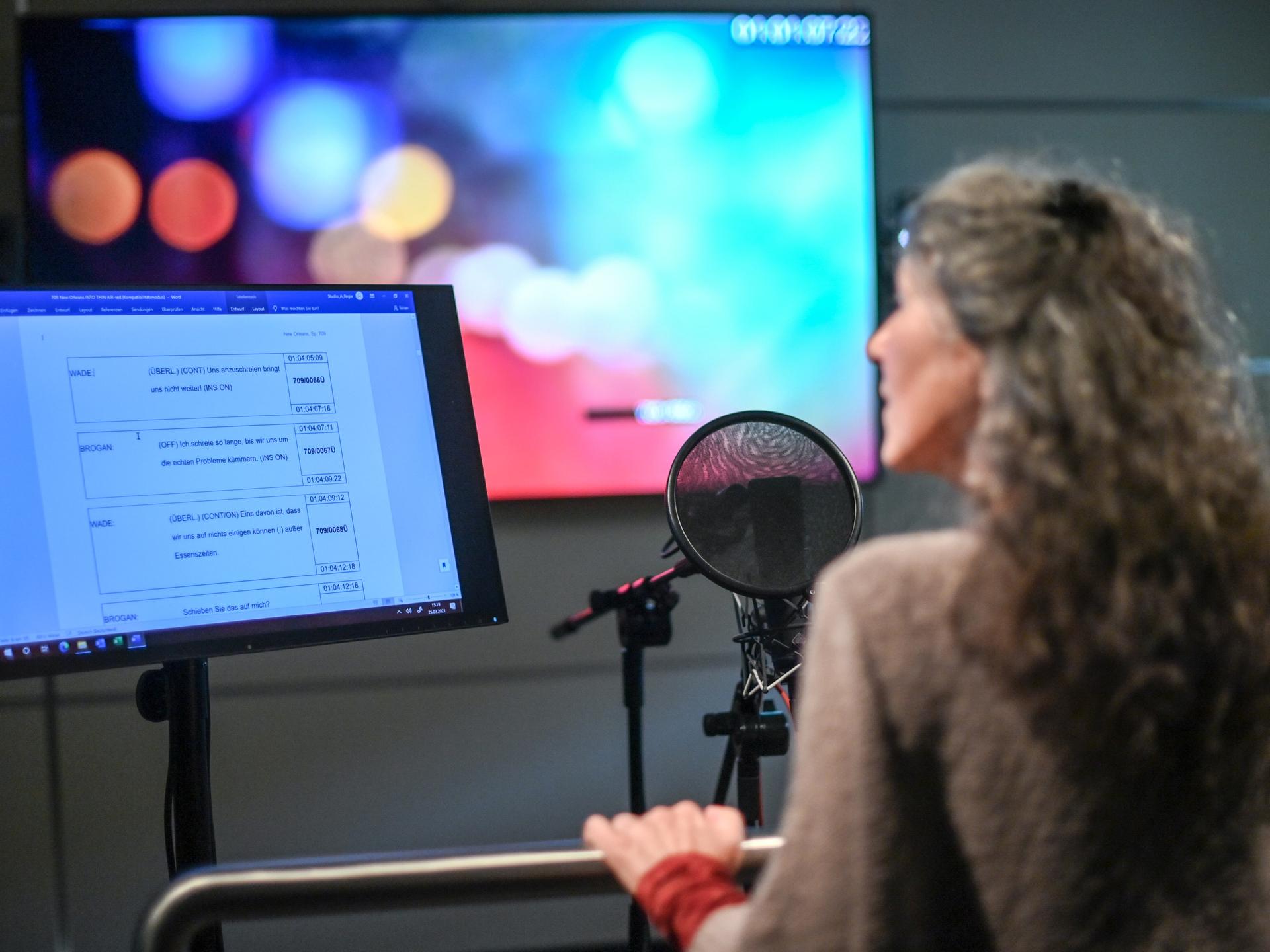 Eine Frau steht in einem Synchronstudio hinter einem Mikrofon und einem Bildschirm, der Dialoge anzeigt.