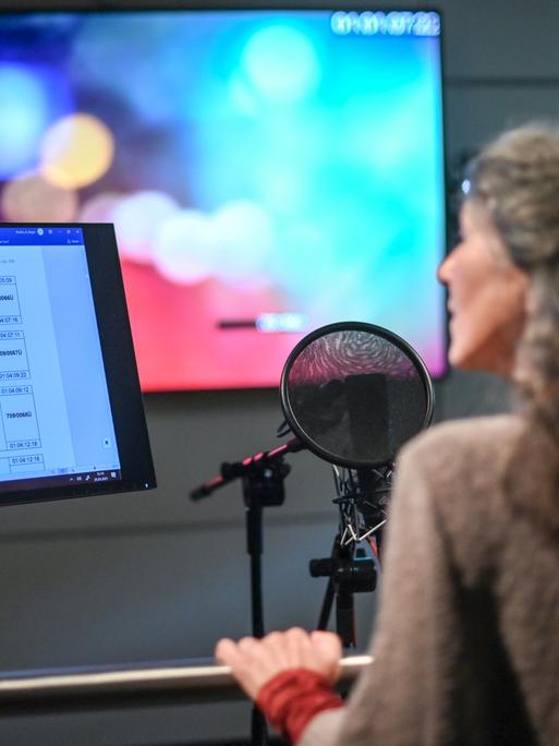 Eine Frau steht in einem Synchronstudio hinter einem Mikrofon und einem Bildschirm, der Dialoge anzeigt.