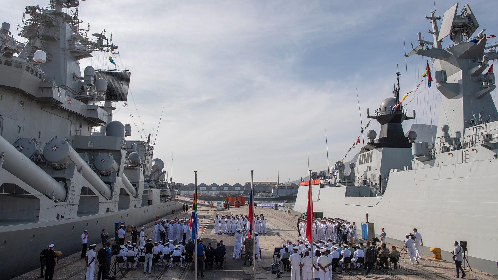 Eröffnung des ersten gemeinsamen Marinemanövers von Südafrika, China und Russland im November 2019 in Kapstadt: zwei Kriegsschiffe, zahlreiche Marinesoldaten und die drei Landesflaggen sind zu sehen.