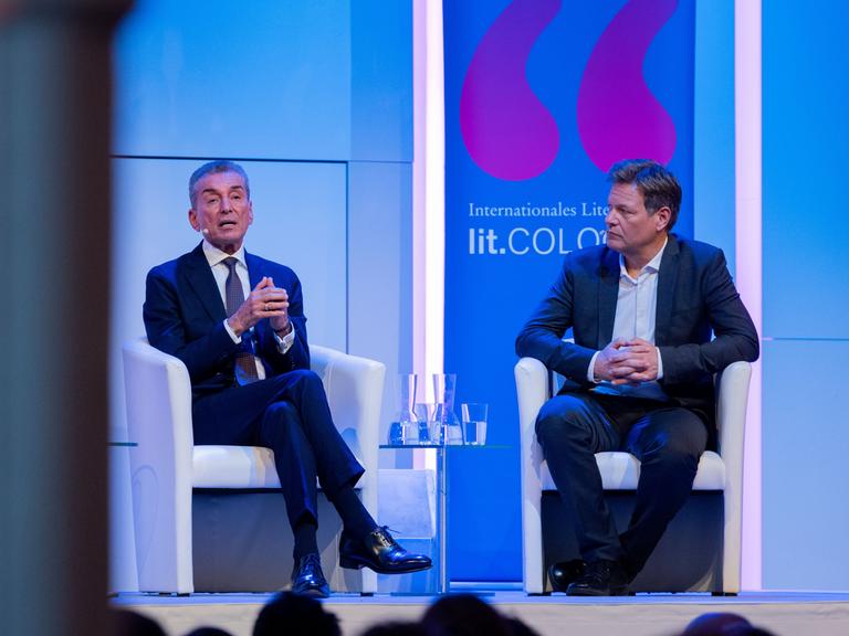 Michel Friedman und Robert Habeck sitzen auf einer Bühne mit blauem Hintergrund. Auf dem Hintergrund ist "Lit.Cologne" zu lesen.