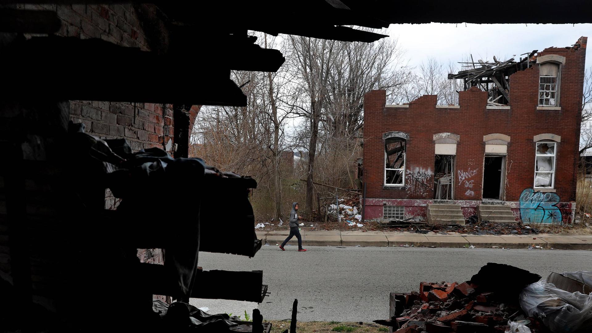 Ein Mann läuft eine Straße entlang. Auf der gegenüberliegenden Straßenseite verfällt ein leerstehendes Wohnhaus. 