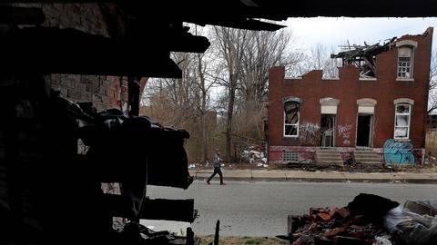 Ein Mann läuft eine Straße entlang. Auf der gegenüberliegenden Straßenseite verfällt ein leerstehendes Wohnhaus. 