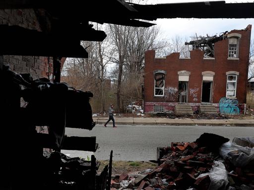Ein Mann läuft eine Straße entlang. Auf der gegenüberliegenden Straßenseite verfällt ein leerstehendes Wohnhaus. 