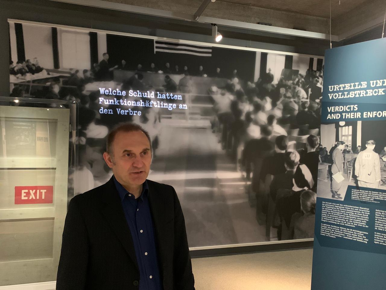 Ein Mann in dunklem Hemd und Jackett steht vor einer großen Fototafel mit einem unvollständigen Schriftzug. Es ist Christoph Thonwald, Leiter der Wissenschaftlichen Abteilung der Gedenkstätte Dachau in Bayern.