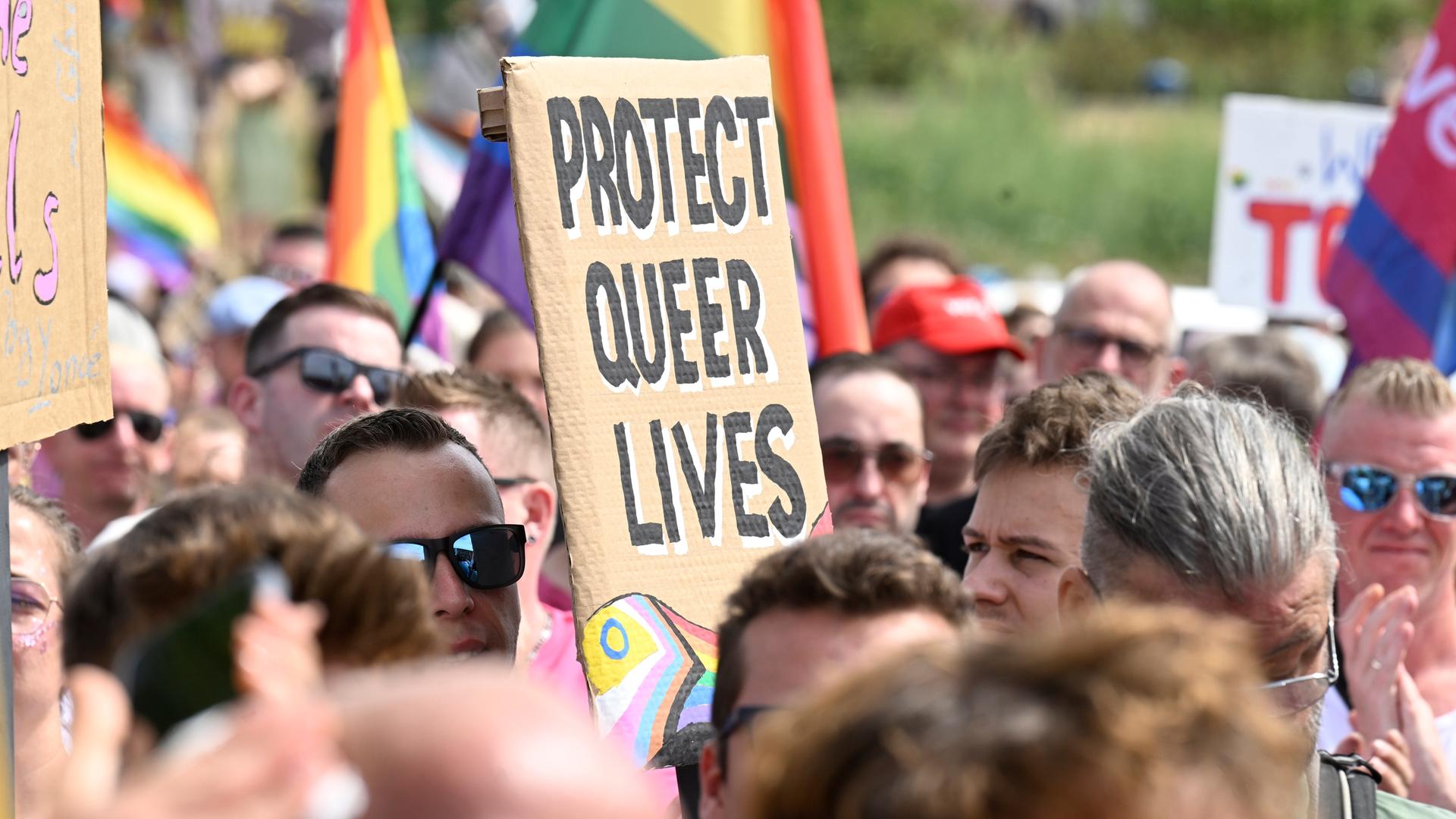Das Bild zeigt ein Protestschild. Es trägt die Aufschrift: "PROTECT QUEER LIVES". Drum herum sind viele Menschen zu sehen.