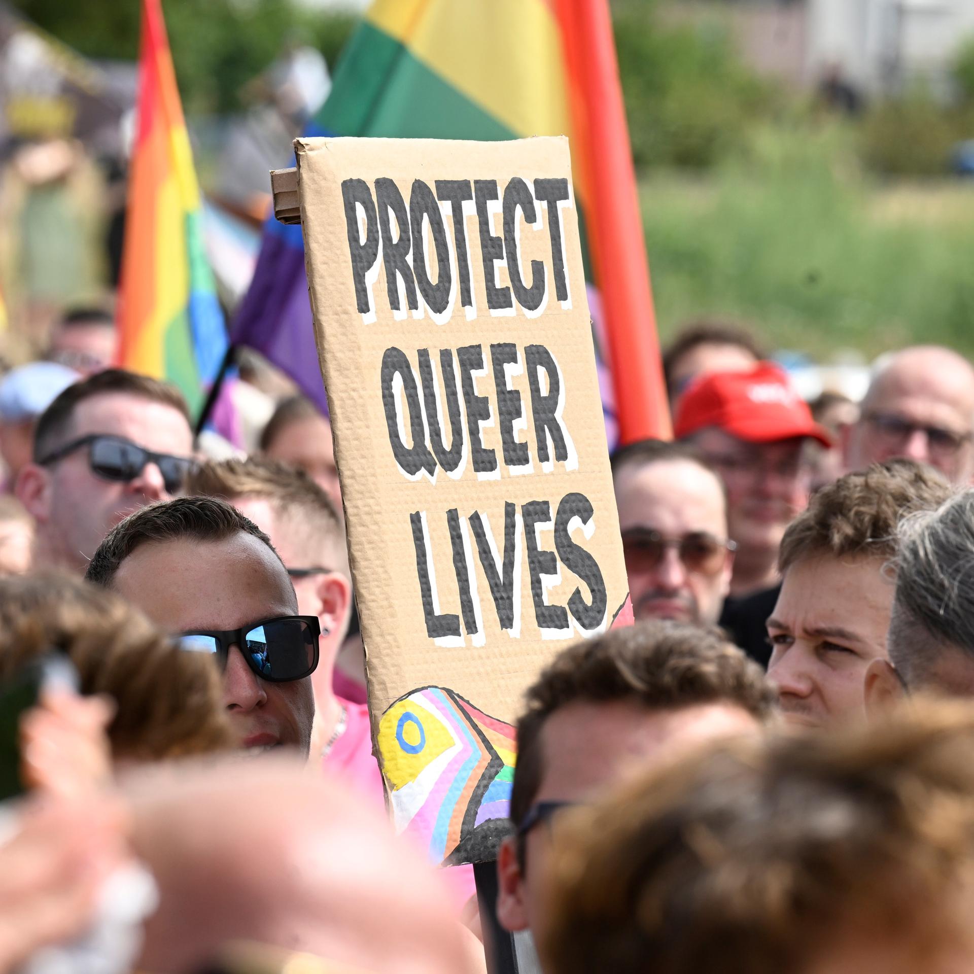Das Bild zeigt ein Protestschild. Es trägt die Aufschrift: "PROTECT QUEER LIVES". Drum herum sind viele Menschen zu sehen.