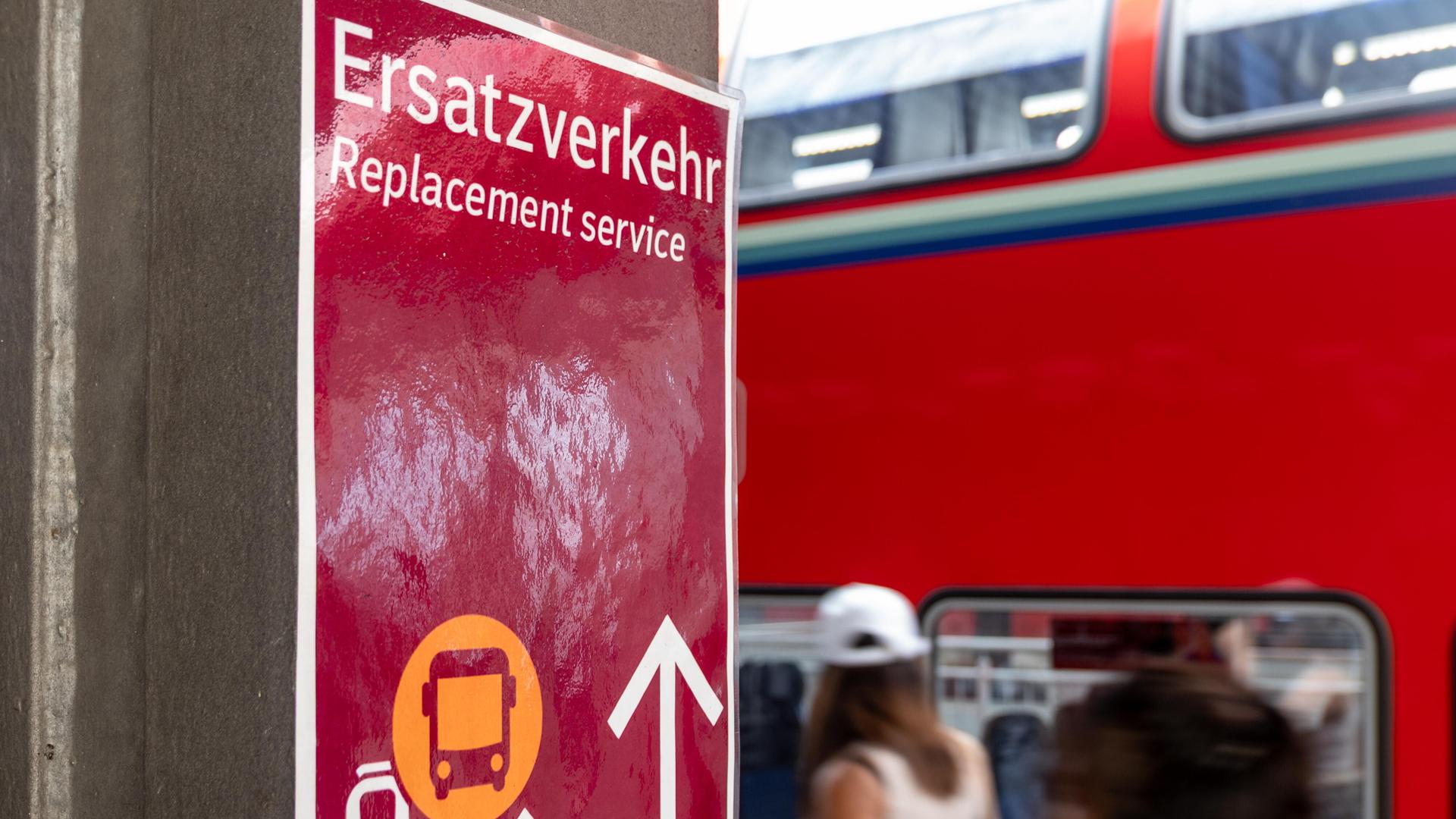 Ein Schild mit dem Hinweis Ersatzverkehr  Replacement service steht gut sichtbar an einem Bahnsteig. Im Hintergrund ist ein doppelstöckiger Regionalzug der Deutschen Bahn (DB) zu sehen.