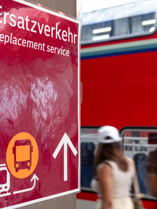 Ein Schild mit dem Hinweis Ersatzverkehr  Replacement service steht gut sichtbar an einem Bahnsteig. Im Hintergrund ist ein doppelstöckiger Regionalzug der Deutschen Bahn (DB) zu sehen.