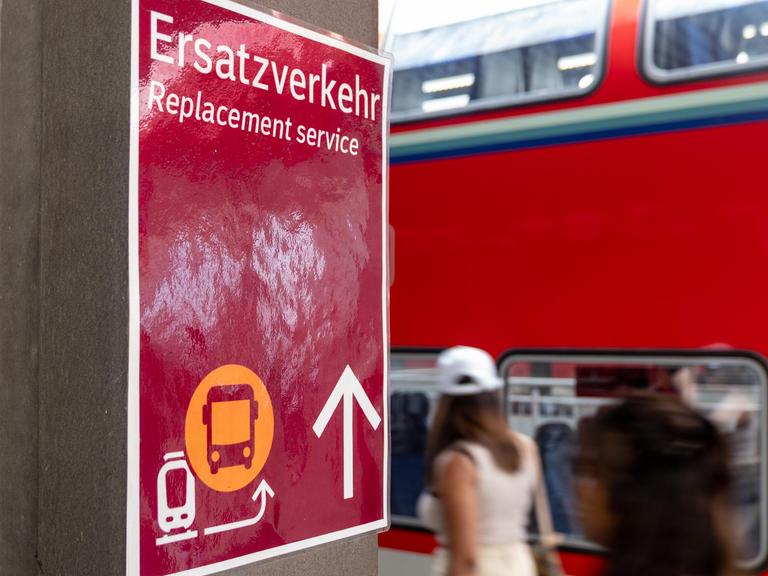 Ein Schild mit dem Hinweis Ersatzverkehr  Replacement service steht gut sichtbar an einem Bahnsteig. Im Hintergrund ist ein doppelstöckiger Regionalzug der Deutschen Bahn (DB) zu sehen.
