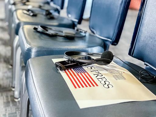 In einem Raum mit vielen Stühlen liegen Kopfhörer und Übersetzungsgeräte auf jedem Stuhl. Auf einem Stuhl liegt ein Zettel mit der US-Flagge und der Aufschrift Press.