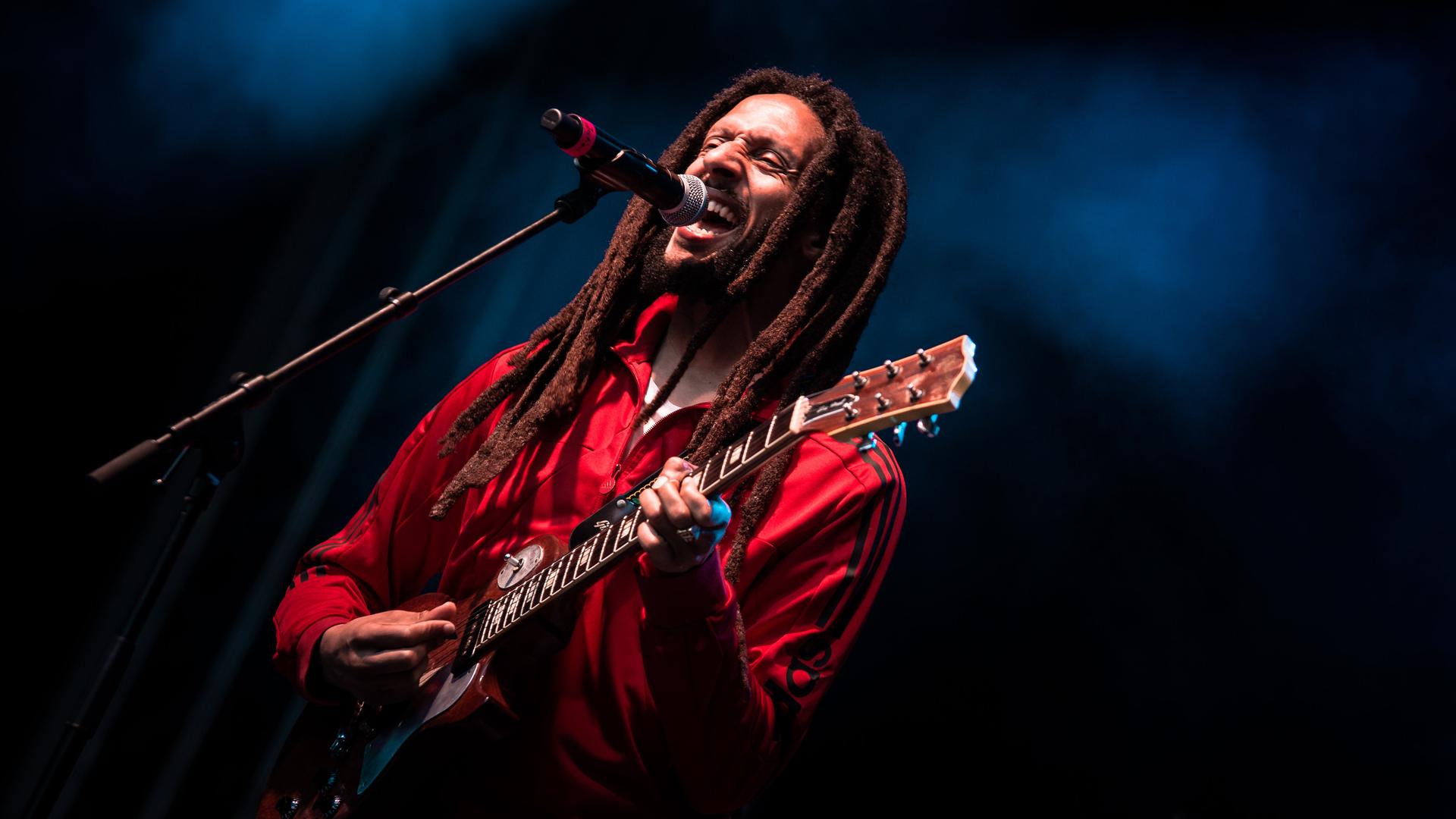 Julian Marley steht mit seiner Gitarre auf der blau ausgeleuchteten Bühne vom Rudolstadt-Festival vor einem Mikrophon und singt.