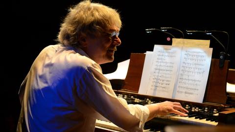 Ein Mann mit kurzen grauen Haaren sitzt vor einem Tasteninstrument mit zwei Manualen. Seine Hände liegen auf den Tasten, er blickt freundlich zur Seite. Vor ihm stehen Notenblätter.