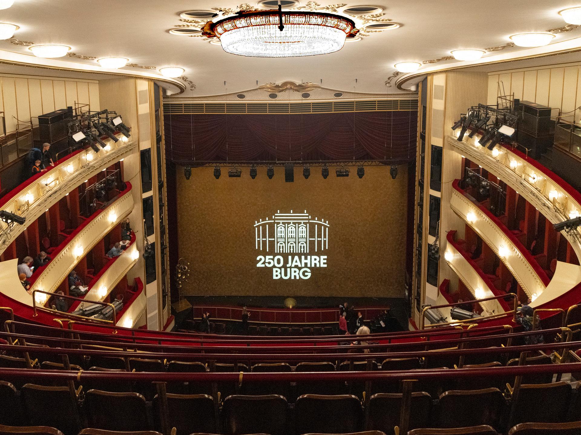 Die Bühne des Wiener Burgtheaters, vom obersten Rang aus gesehen. Zwischen dem roten Vorhang auf der Bühne steht auf einer Leinwand: "250 Jahre Burg"