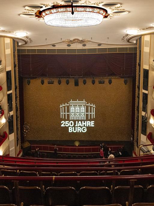 Die Bühne des Wiener Burgtheaters, vom obersten Rang aus gesehen. Zwischen dem roten Vorhang auf der Bühne steht auf einer Leinwand: "250 Jahre Burg"