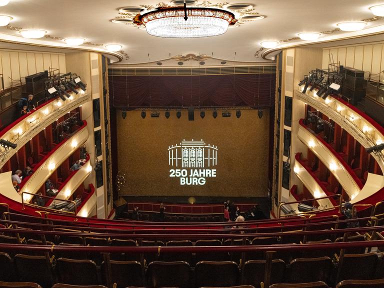 Die Bühne des Wiener Burgtheaters, vom obersten Rang aus gesehen. Zwischen dem roten Vorhang auf der Bühne steht auf einer Leinwand: "250 Jahre Burg"
