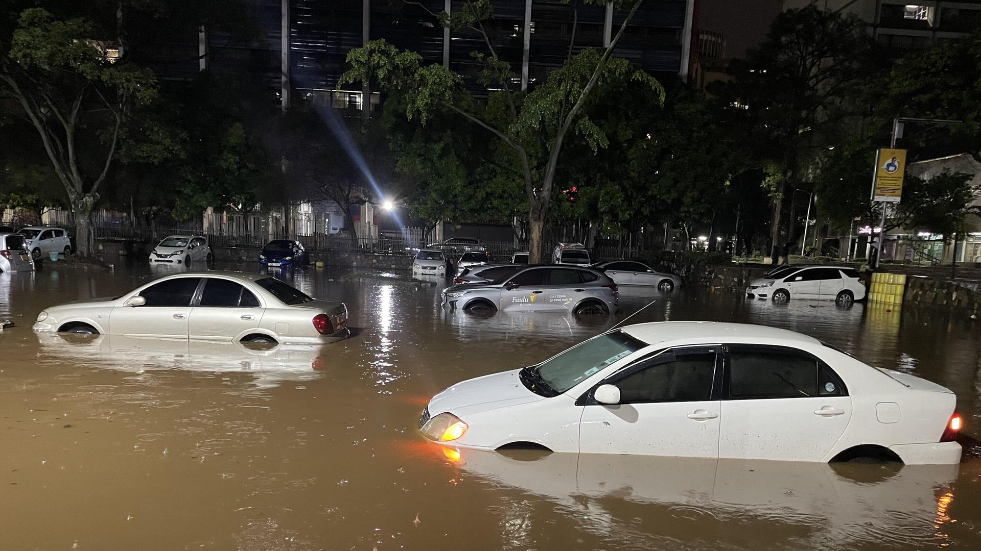 Mehrere Autos stehen bis über die Reifen in braunem Wasser, im Hintergrund sind einige Bäume zu sehen.