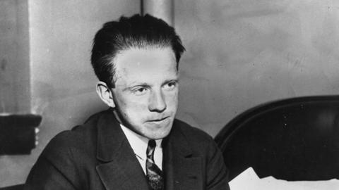Werner Heisenberg schaut in den Raum, vor ihm auf dem Schreibtisch liegt ein Buch oder ein Notizbuch