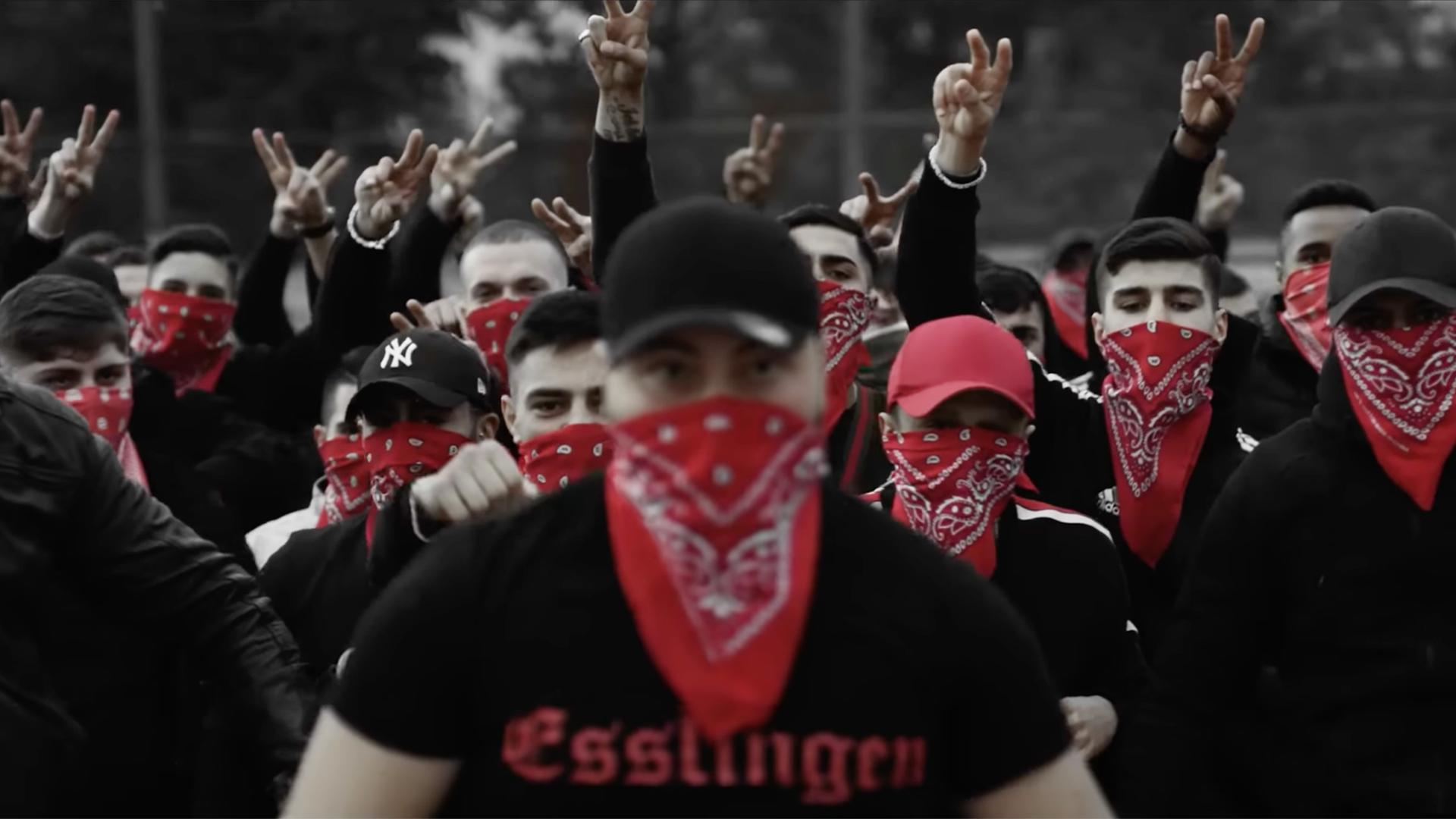 Screenshot aus dem Musikvideo "Eska - Bumaye". Eine Gruppe vermummter junger Männer mit der Aufschrift "Esslingen" auf schwarzer Kleidung. Rapper Eska wird mit einer Bande in Verbindung gebracht. In seinen Musikvideos inszenieren sich Männer als Gangster.