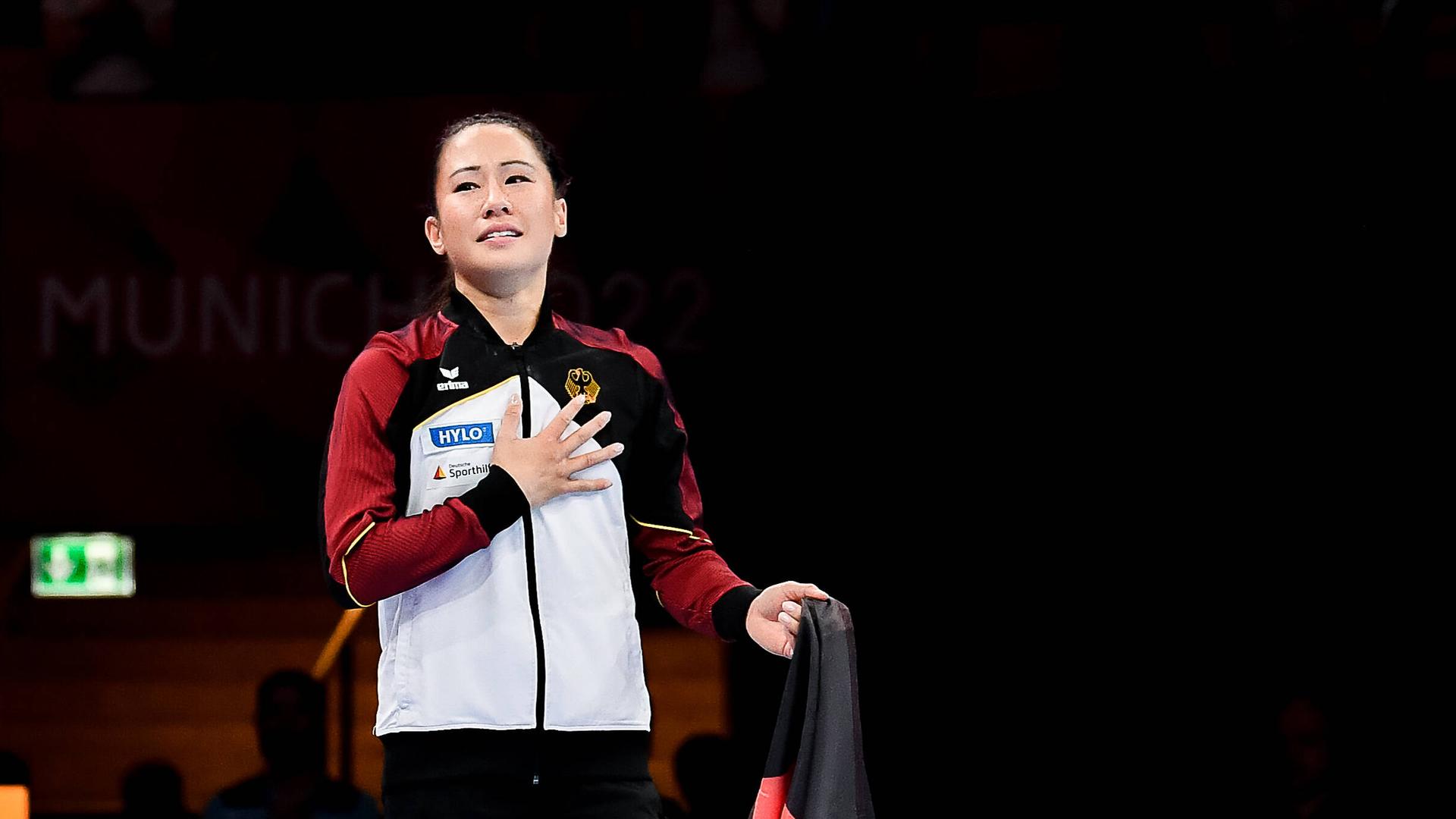 Kim Bui beendet ihre Karriere als Kunstturnerin und verabschiedet sich von der Öffentlichkeit nach dem Finale im Stufenbarren bei den Europameisterschaften in München 2022. Zu sehen ist sie im Turnanzug der deutschen Nationalmannschaft mit einer Flagge und sichtlich berührt. Kim Bui beendet ihre Karriere als Kunstturnerin und verabschiedet sich von der Öffentlichkeit nach dem Finale im Stufenbarren bei den Europameisterschaften in München 2022. Zu sehen ist sie im Turnanzug der deutschen Nationalmannschaft mit einer Flagge und sichtlich berührt.