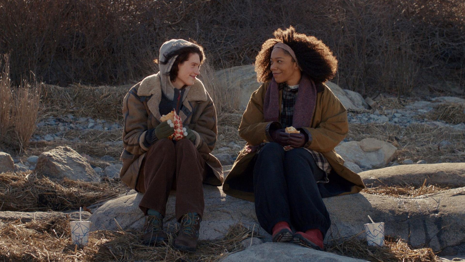 Szene aus dem Film "Sorry Baby": Die Freundinnen Agnes (Eva Victor) und Lydie (Naomie Ackie) sitzen in der Natur auf größeren Steinen und lächeln sich an. Szene aus dem Film "Sorry Baby": Die Freundinnen Agnes (Eva Victor) und Lydie (Naomie Ackie) sitzen in der Natur auf größeren Steinen und lächeln sich an.