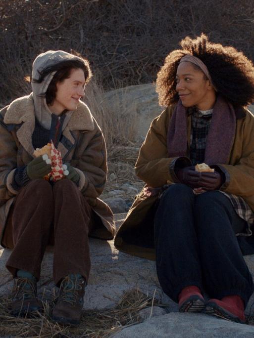 Szene aus dem Film "Sorry Baby": Die Freundinnen Agnes (Eva Victor) und Lydie (Naomie Ackie) sitzen in der Natur auf größeren Steinen und lächeln sich an.