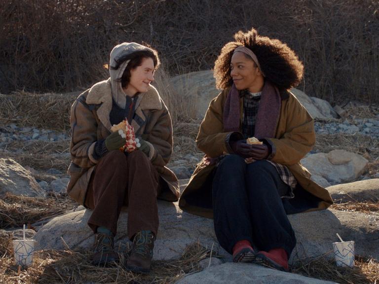 Szene aus dem Film "Sorry Baby": Die Freundinnen Agnes (Eva Victor) und Lydie (Naomie Ackie) sitzen in der Natur auf größeren Steinen und lächeln sich an.