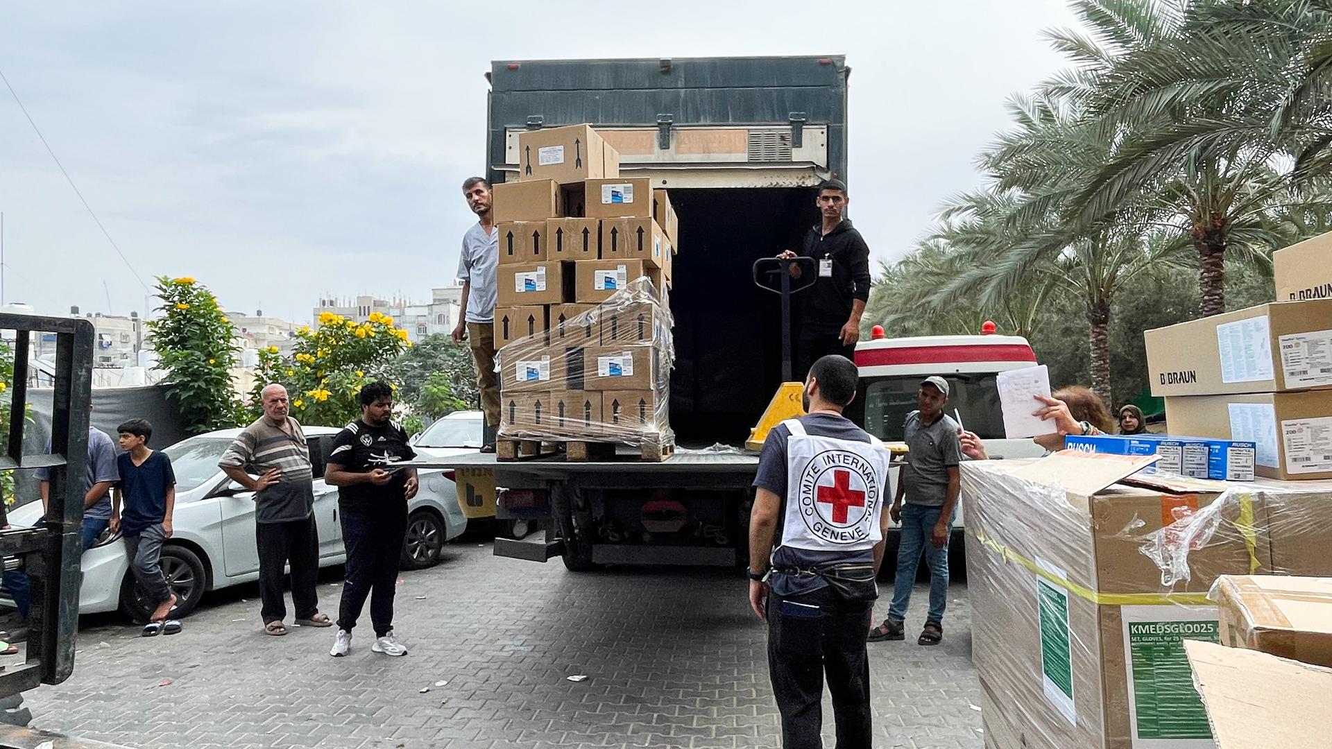 Mehrere Menschen stehen um einen LKW herum, der auf seiner Ladefläche viele Paketen hat. Auf dem Boden stehen mehrere Pakete. Im Vordergrund steht neben einem Gabelstapler ein Mann mit einer Weste mit dem Symbol des Internationalen Roten Kreuzes. Im Hintergrund sind Palmen und Häuser.
