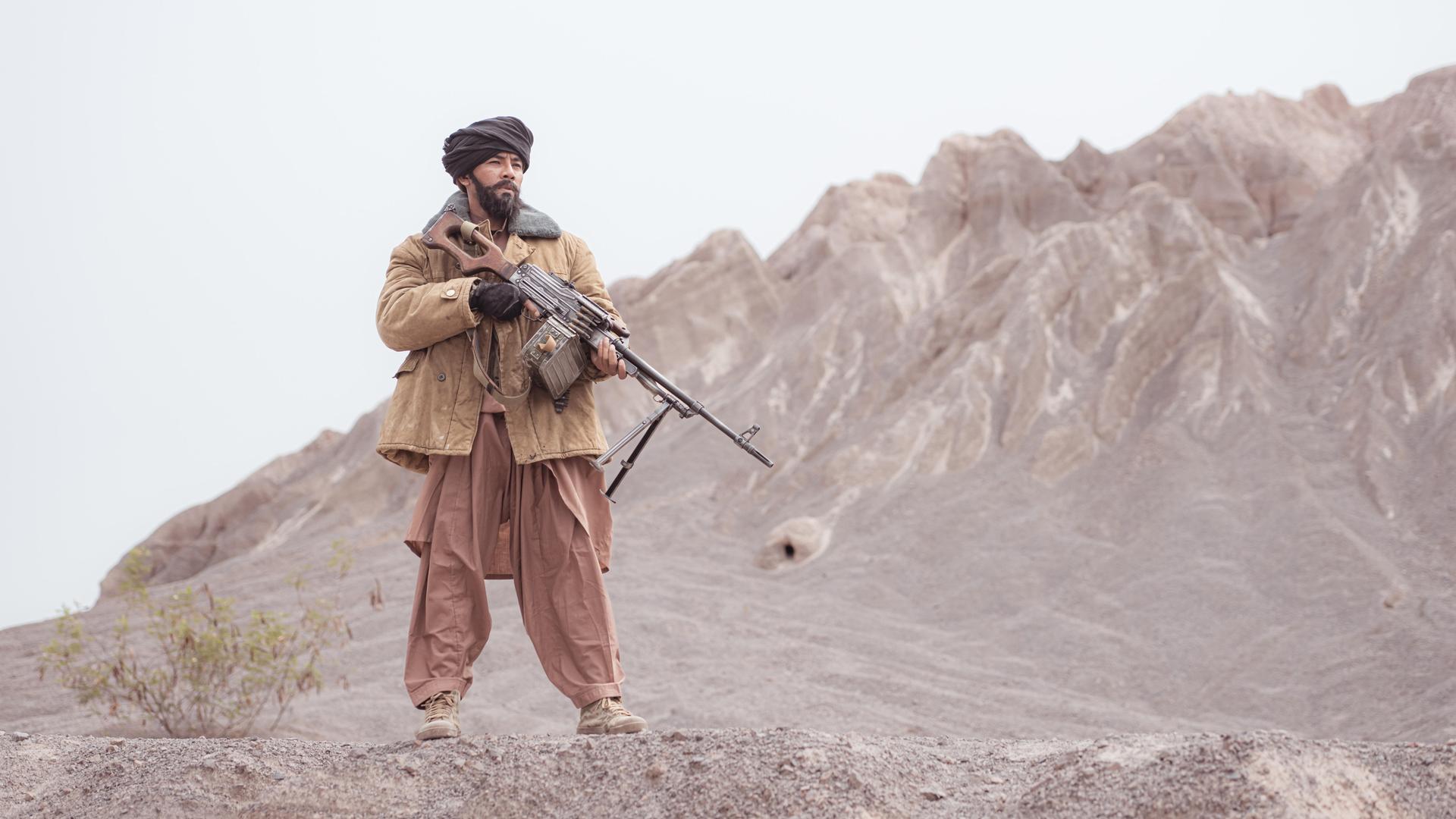 Ein Kämpfer der Taliban steht mit einem Maschinengewehr in der Wüste, im Hintergrund sieht man Berge.