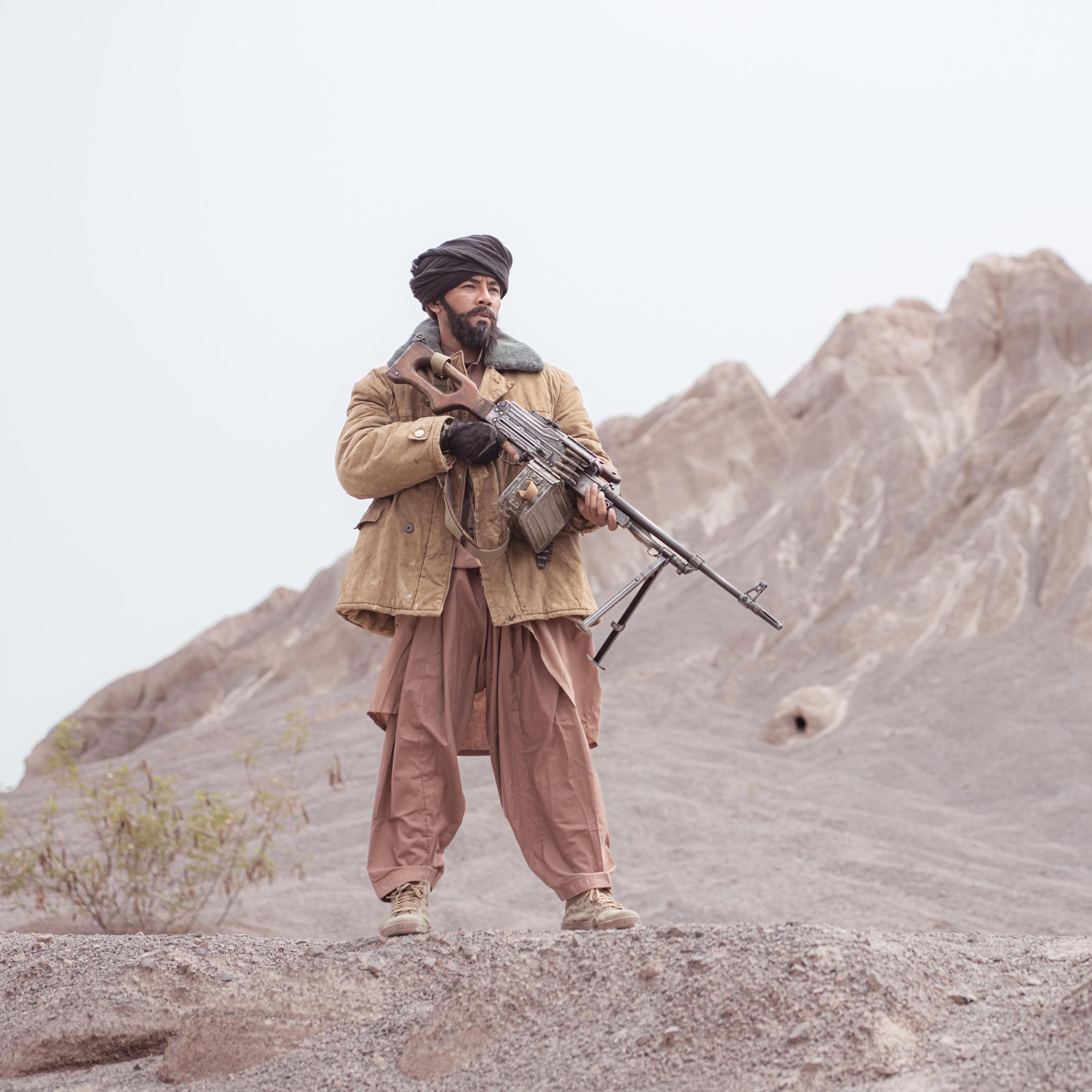 Ein Kämpfer der Taliban steht mit einem Maschinengewehr in der Wüste, im Hintergrund sieht man Berge.