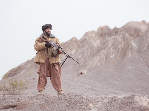 Ein Kämpfer der Taliban steht mit einem Maschinengewehr in der Wüste, im Hintergrund sieht man Berge.