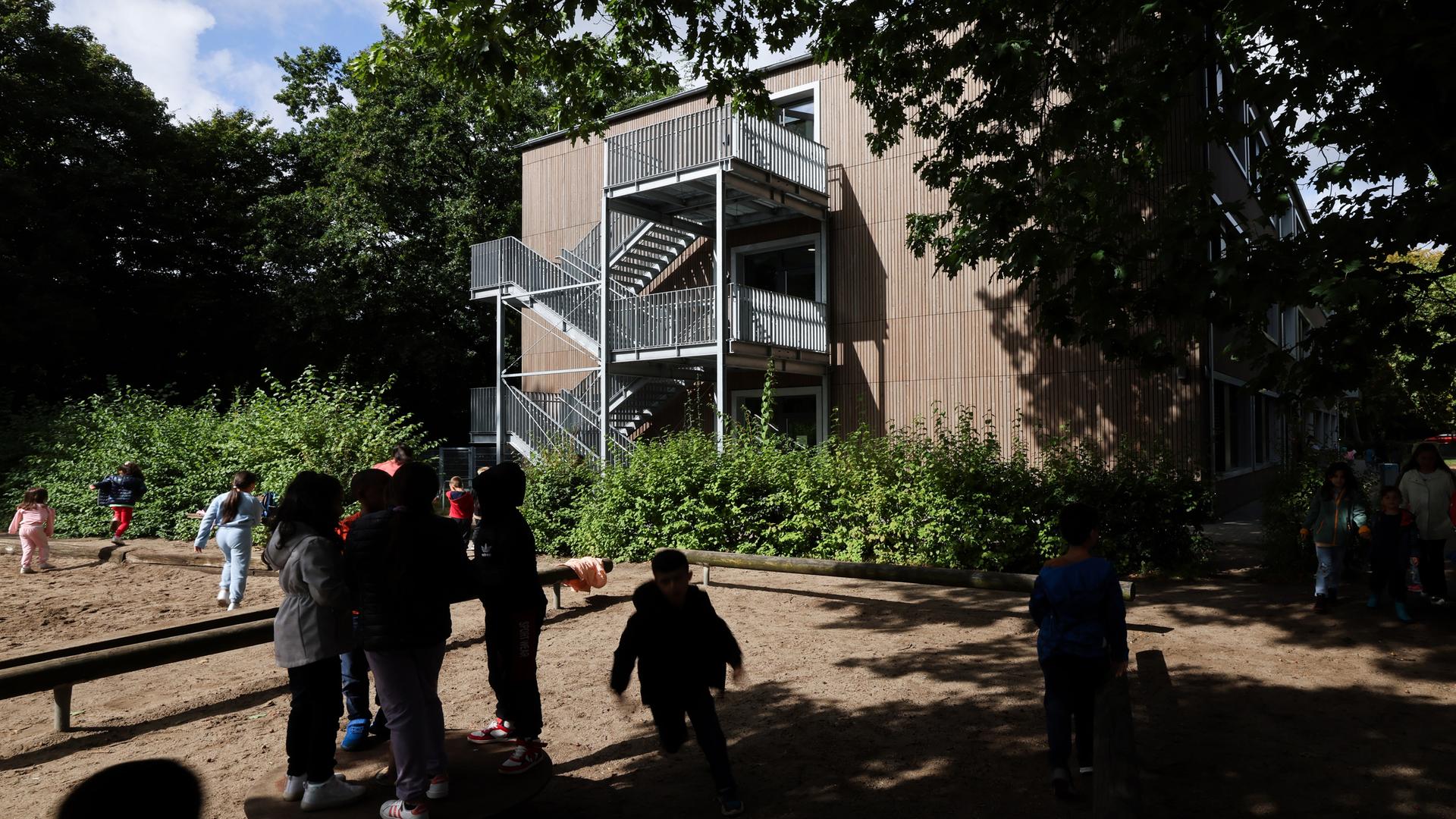 Blick auf ein Schulgebäude. Davor spielen Kinder im Schatten der Bäume.