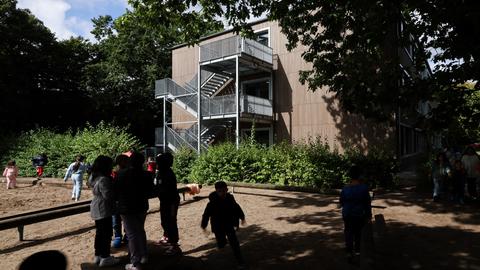 Blick auf ein Schulgebäude. Davor spielen Kinder im Schatten der Bäume.