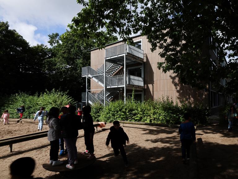 Blick auf ein Schulgebäude. Davor spielen Kinder im Schatten der Bäume.