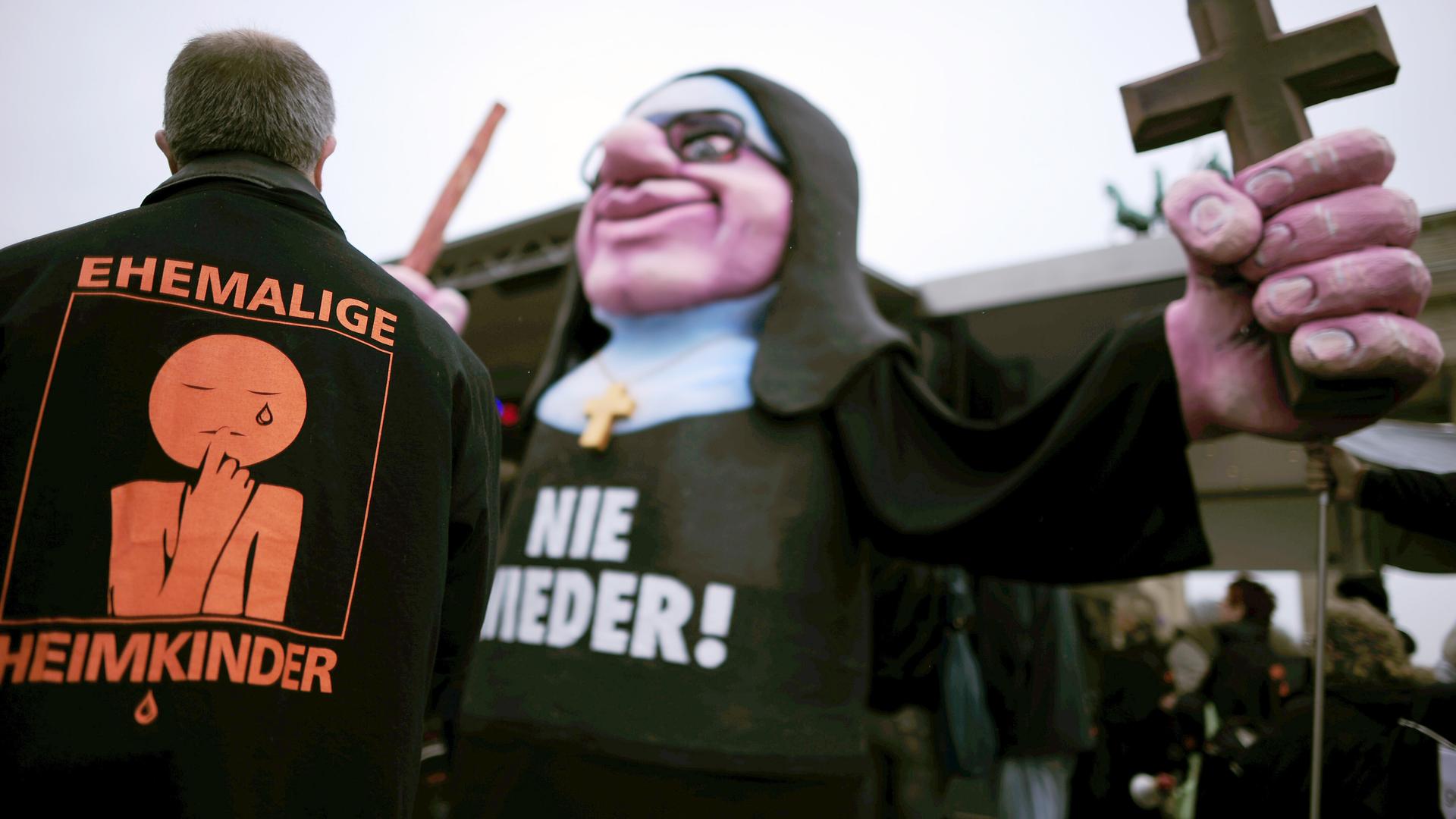 Ein Mann trägt auf dem Rücken die orangefarbene Aufschrift des Vereins "Ehemalige Heimkinder". Daneben ist eine Nonna als lebensgroße Pappmaché-Puppe zu sehen, sie hält ein Kreuz in einer überdimensionalen Hand. Auf ihrem Bauch steht "Nie wieder". 