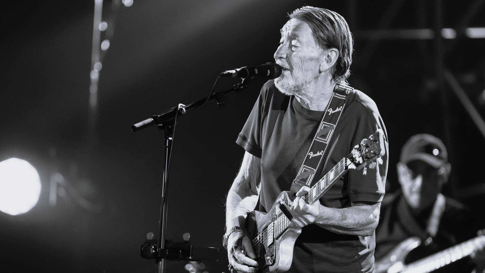 Chris Rea steht auf der Bühne. Er spielt Gitarre und singt in ein Mikrofon. Das Bild ist schwarz-weiß.