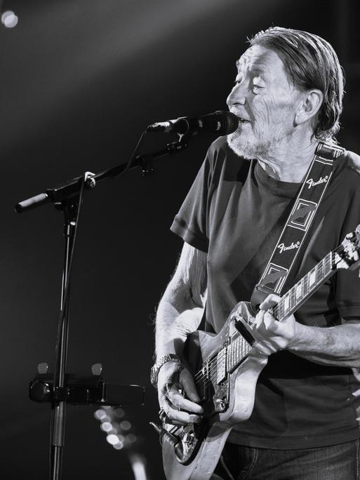 Chris Rea steht auf der Bühne. Er spielt Gitarre und singt in ein Mikrofon. Das Bild ist schwarz-weiß. (Quelle: picture alliance)